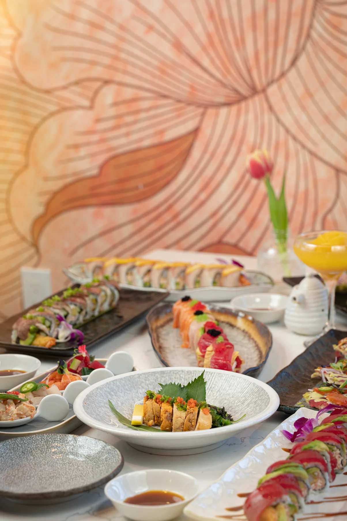 A styled table spread of sushi rolls, sashimi, and a cocktail at Fish Boon, with the hand-painted crane mural behind.